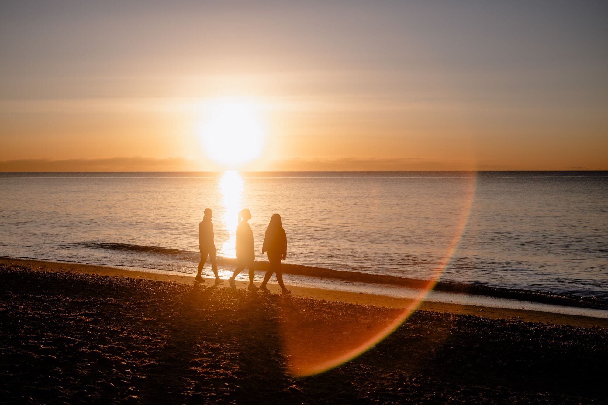 Un atardecer sobre una playa con tres personas silueteadas contra el sol.