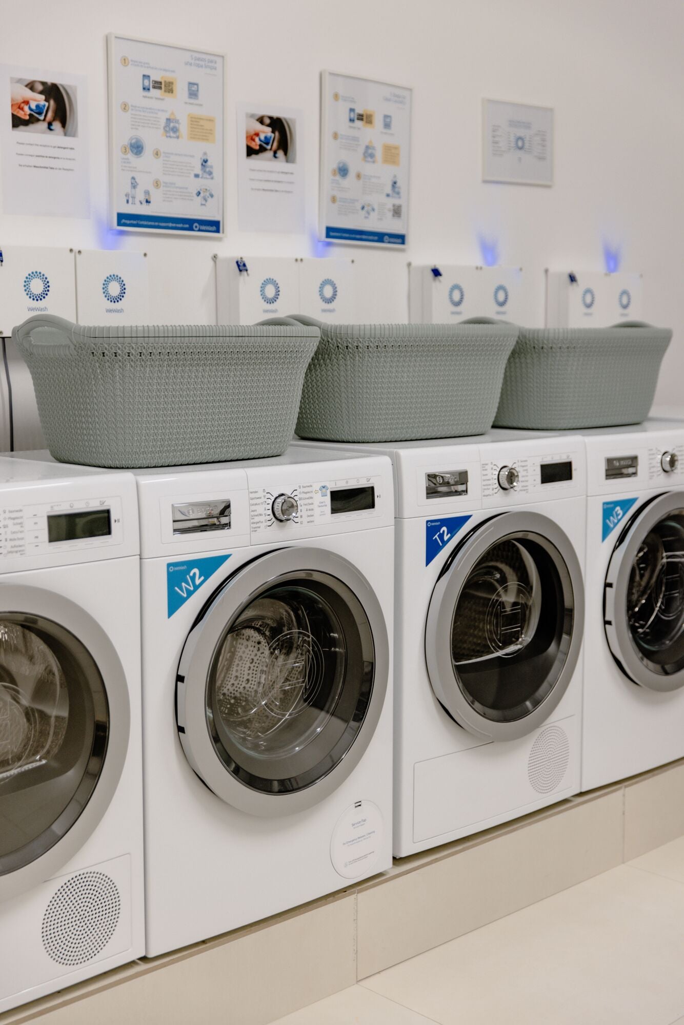 A row of front-loading washing machines with baskets on top, arranged in a commercial setting.