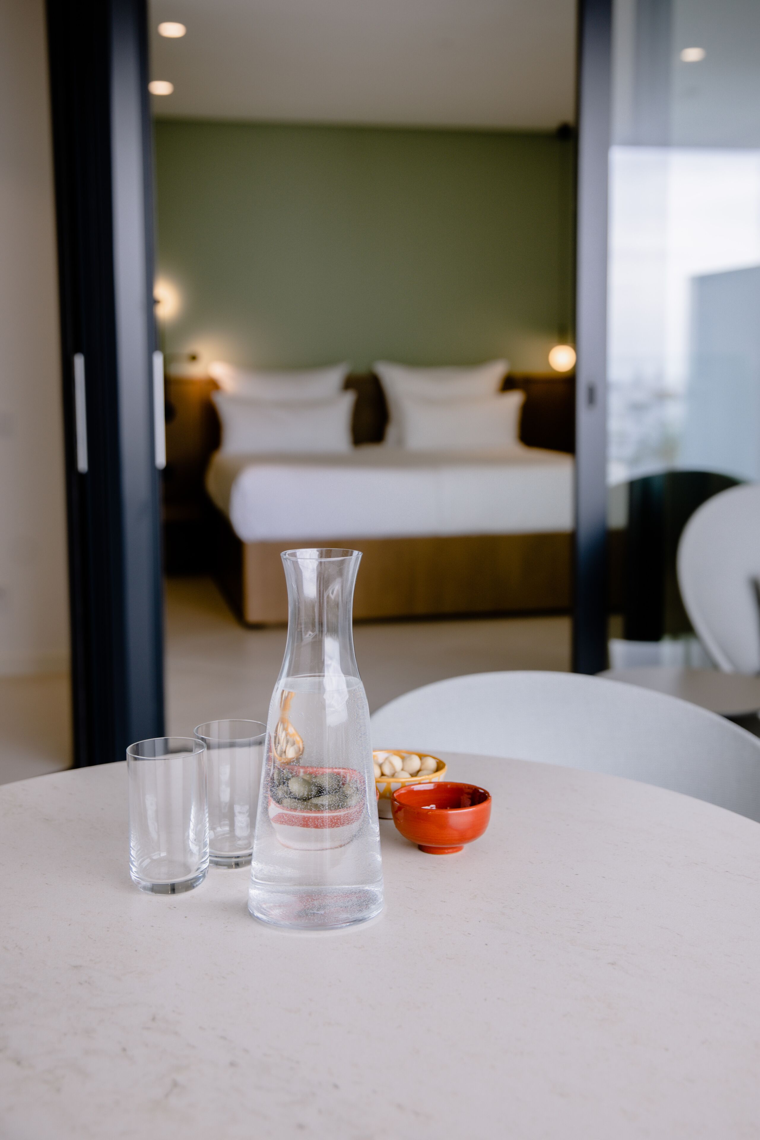 Una habitación de hotel con una cama, una mesita de noche con un florero, vasos y un cuenco.
