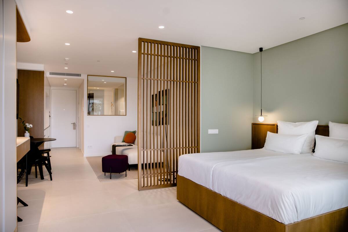 A modern hotel room with a large bed, a desk with a chair, a small table, and a partition.