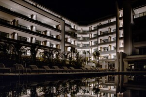 A multi-story hotel with balconies, a pool area with lounge chairs, and palm trees.