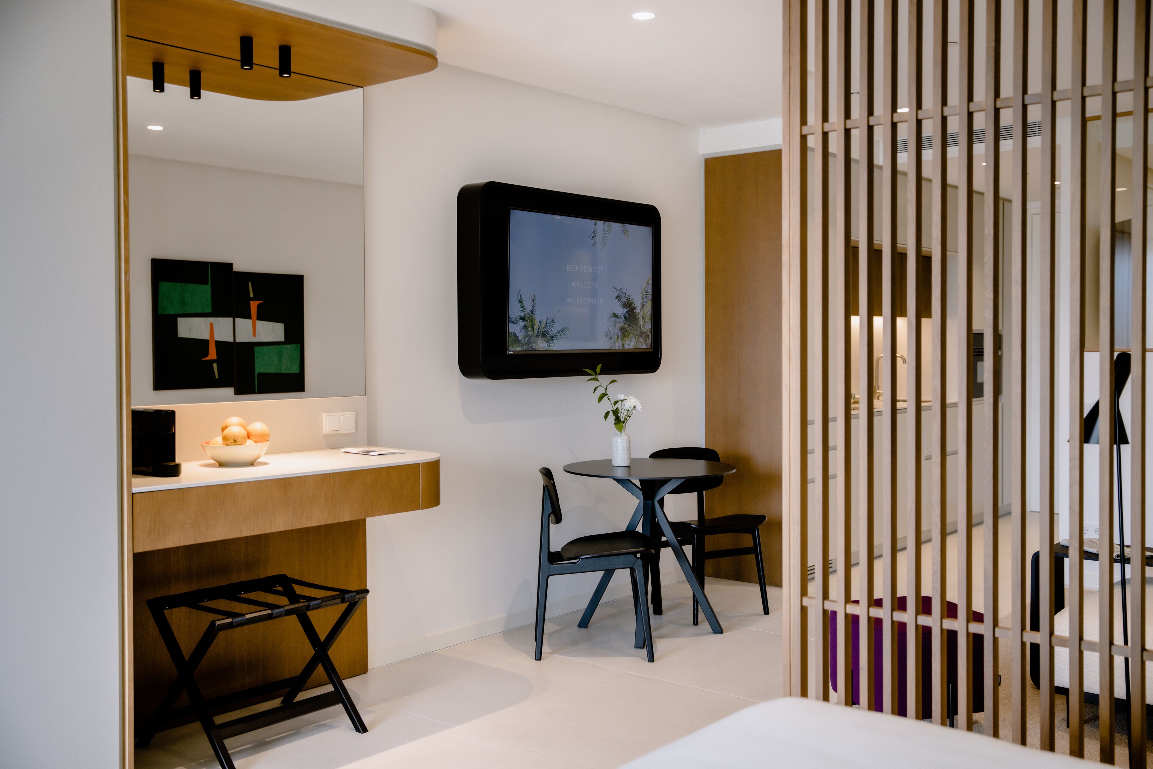 Una habitación de hotel moderna con un televisor de pantalla plana montado en la pared, un escritorio de madera con un frutero, dos sillas y una mesita con un jarrón de flores.