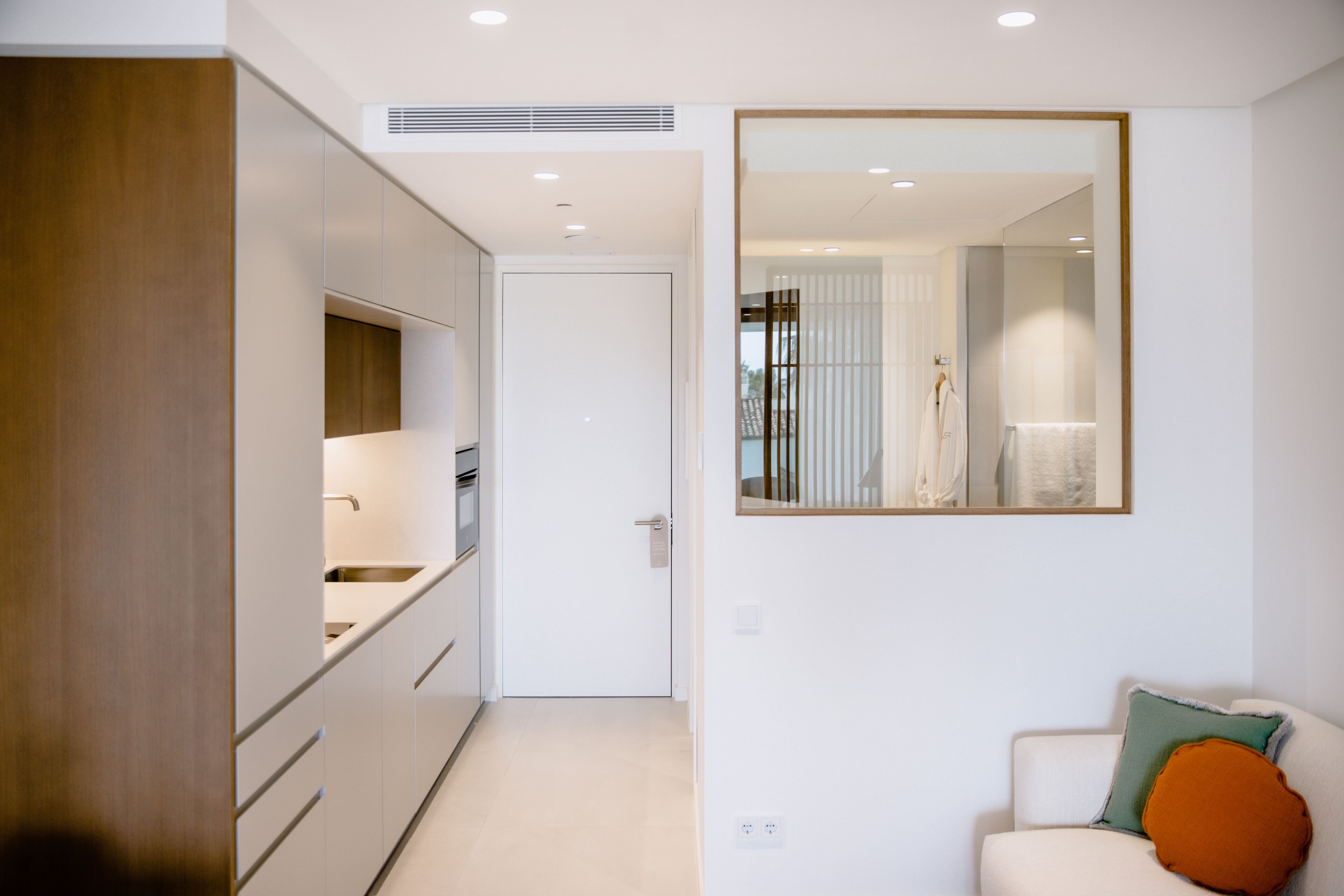 A modern kitchen with white cabinets, a built-in oven, and a sink. There is a white door leading to another room. A large mirror hangs on the wall above a white sofa with an orange pillow.