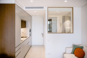 A modern kitchen with white cabinets, a built-in oven, and a sink. There is a white door leading to another room. A large mirror hangs on the wall above a white sofa with an orange pillow.