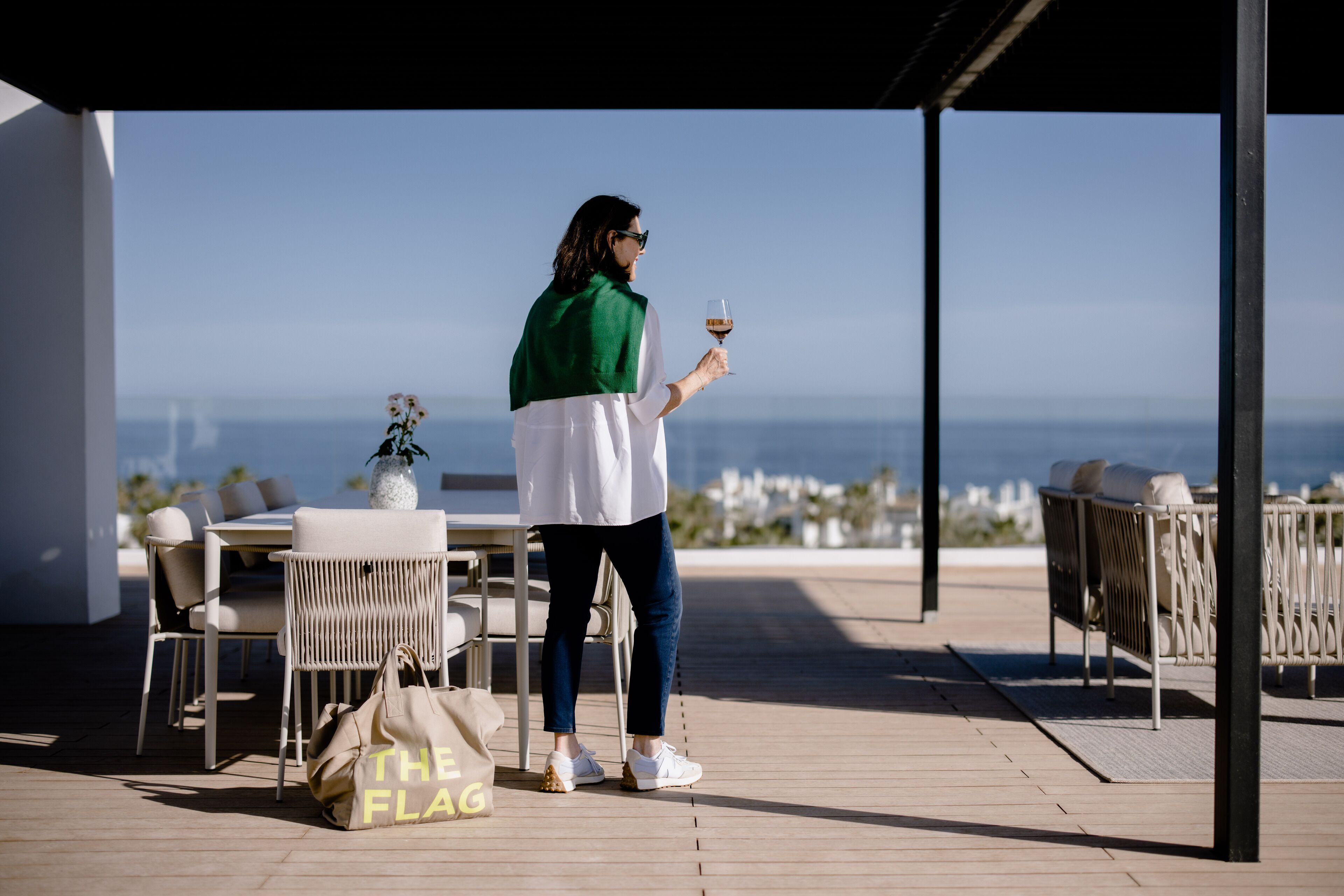 A person standing on a wooden deck holding a glass of wine, with a view of the ocean and a table with chairs in the background.