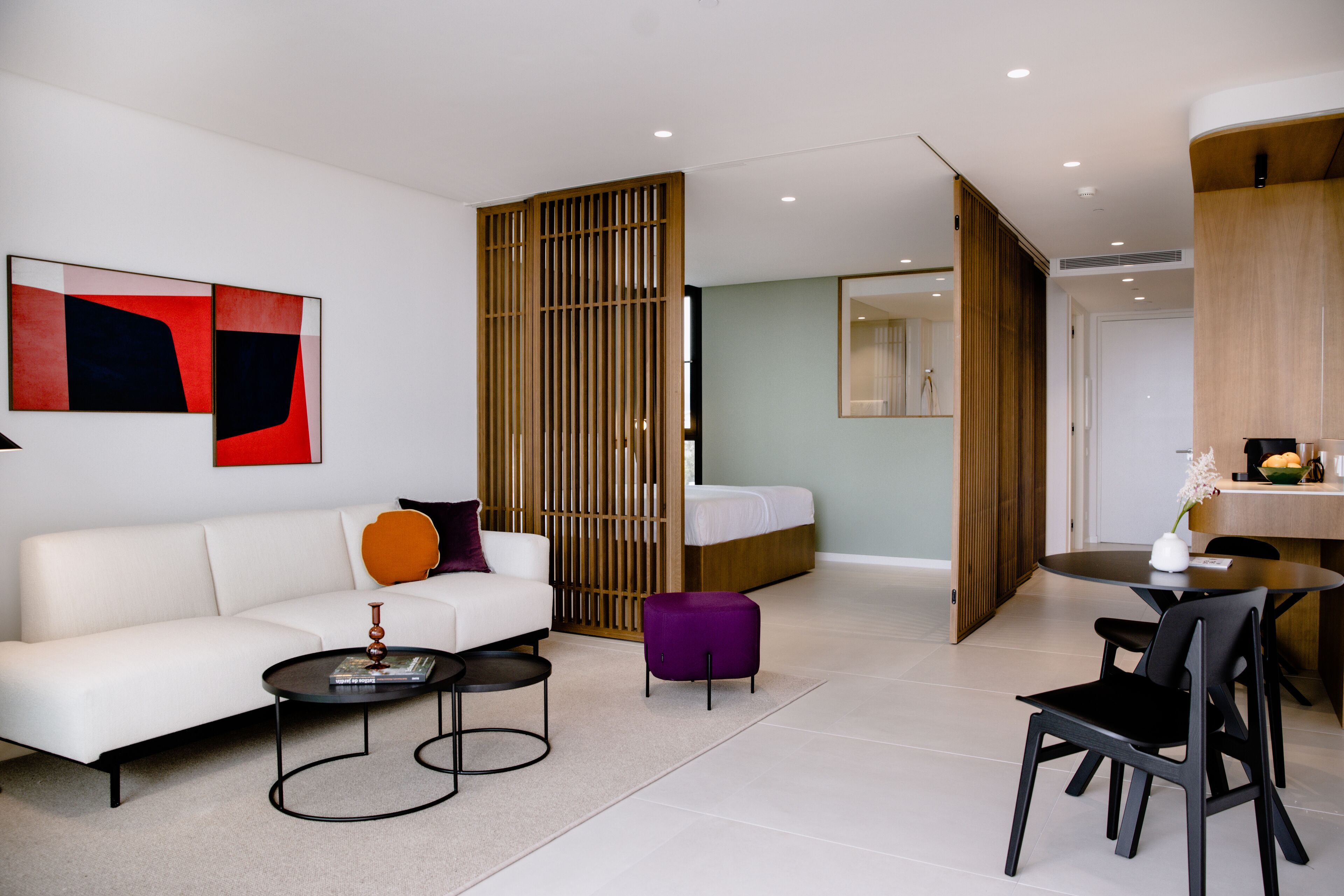 Una sala moderna con un sofá blanco, una mesa de centro y un reposapiés morado. Se observa una partición de madera y una cama al fondo.