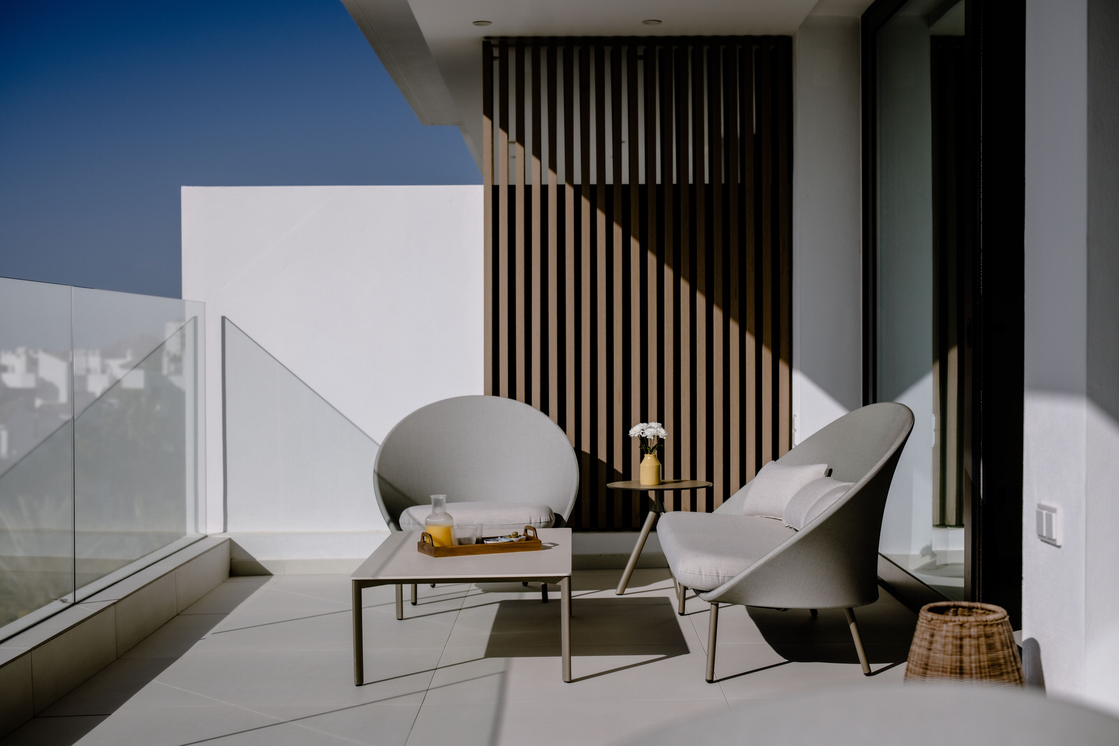 A modern balcony with a glass railing, a white table with a tray, two grey lounge chairs, and a small round table with a vase of flowers.