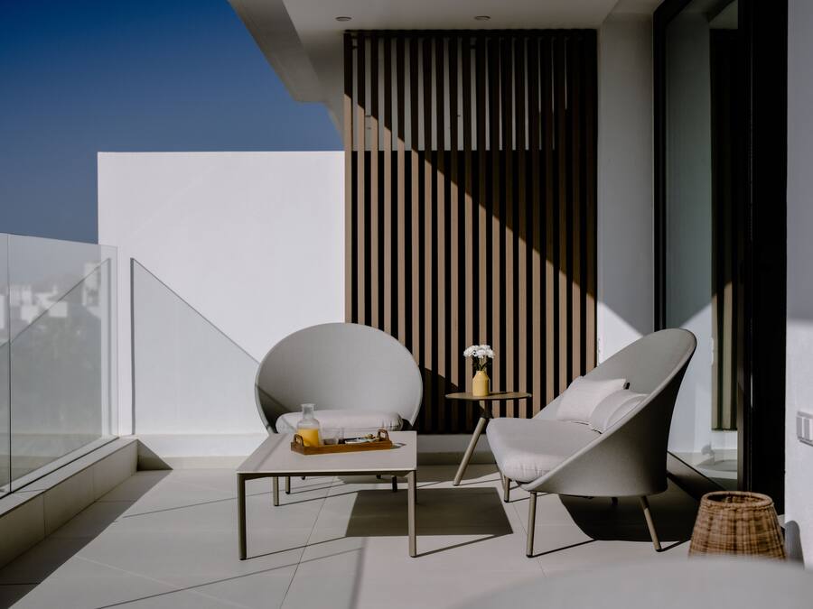 A modern balcony with a glass railing, a white table with a tray, two grey lounge chairs, and a small round table with a vase of flowers.