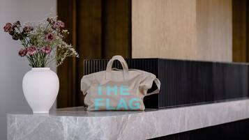 Un jarrón blanco con flores y un bolso color beige con el texto "THE FLAG" sobre una repisa de mármol.