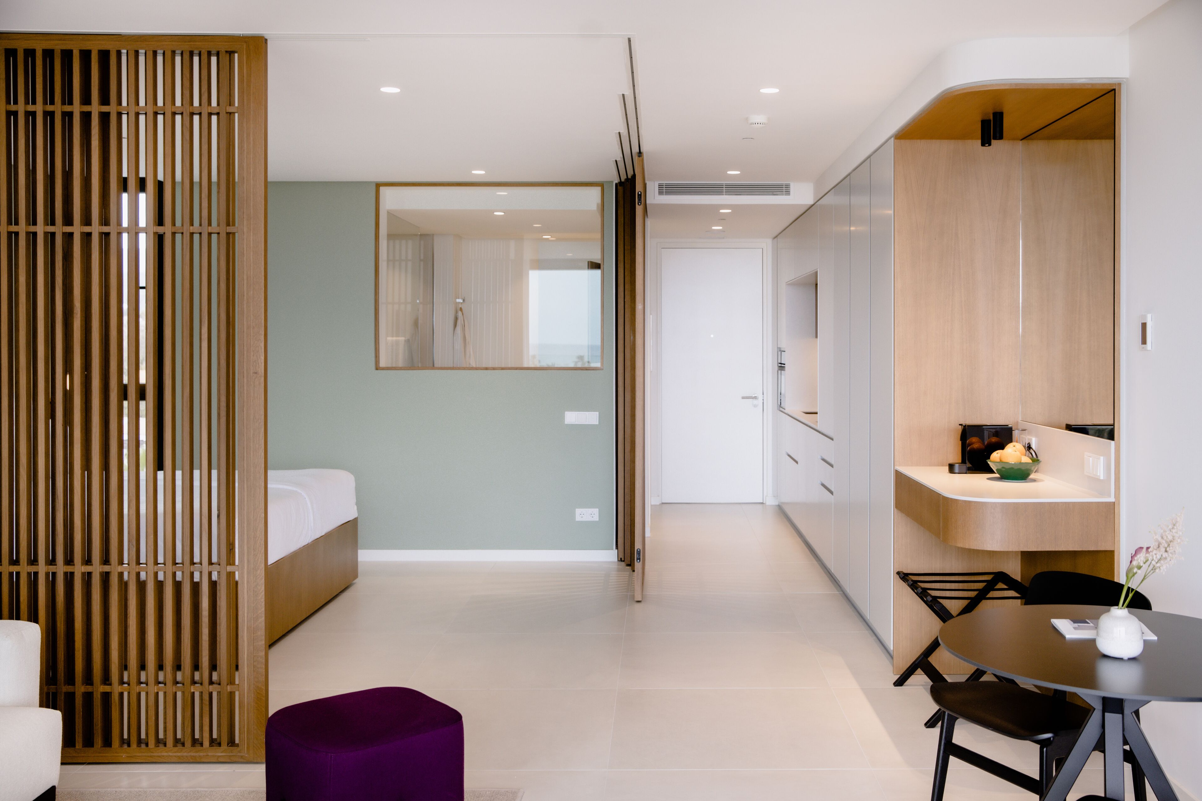 Una habitación de hotel moderna con cama, un separador de madera, un área de comedor con mesa y sillas, y una cocina con armarios y un lavamanos.