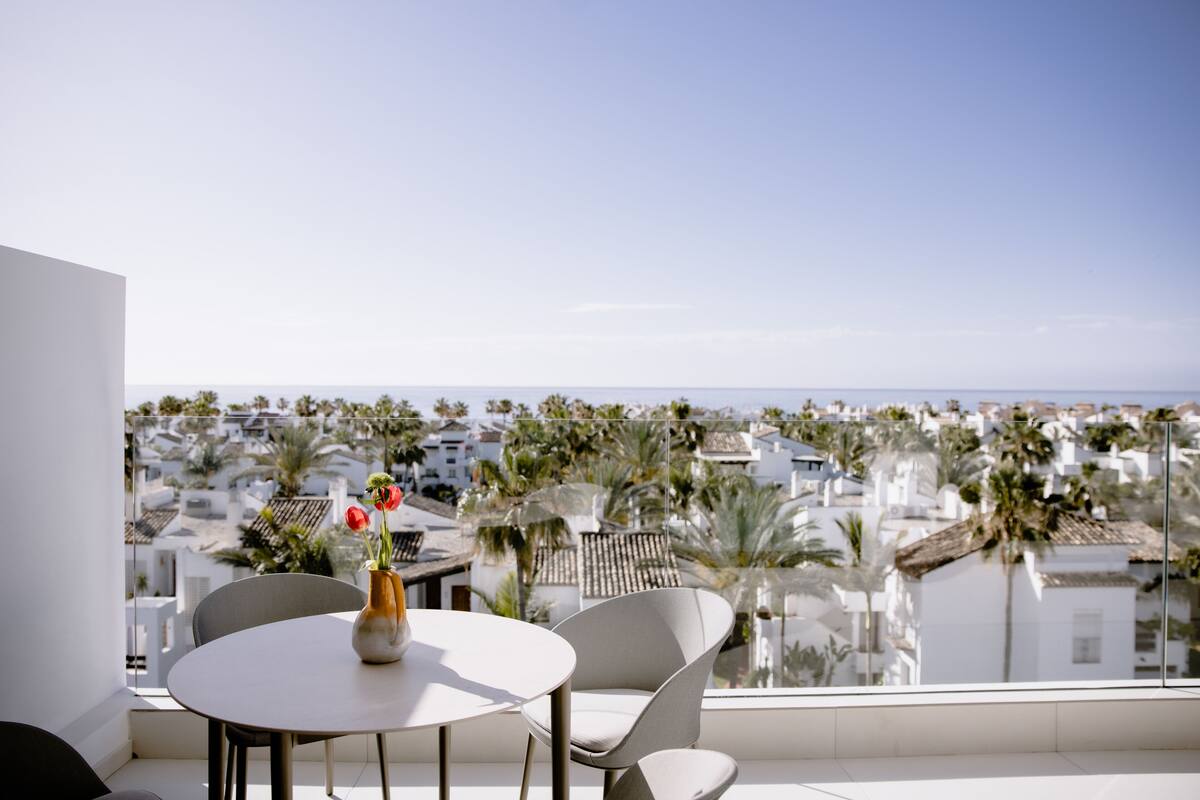 A balcony with a table and chairs, overlooking a coastal town with palm trees and white buildings.