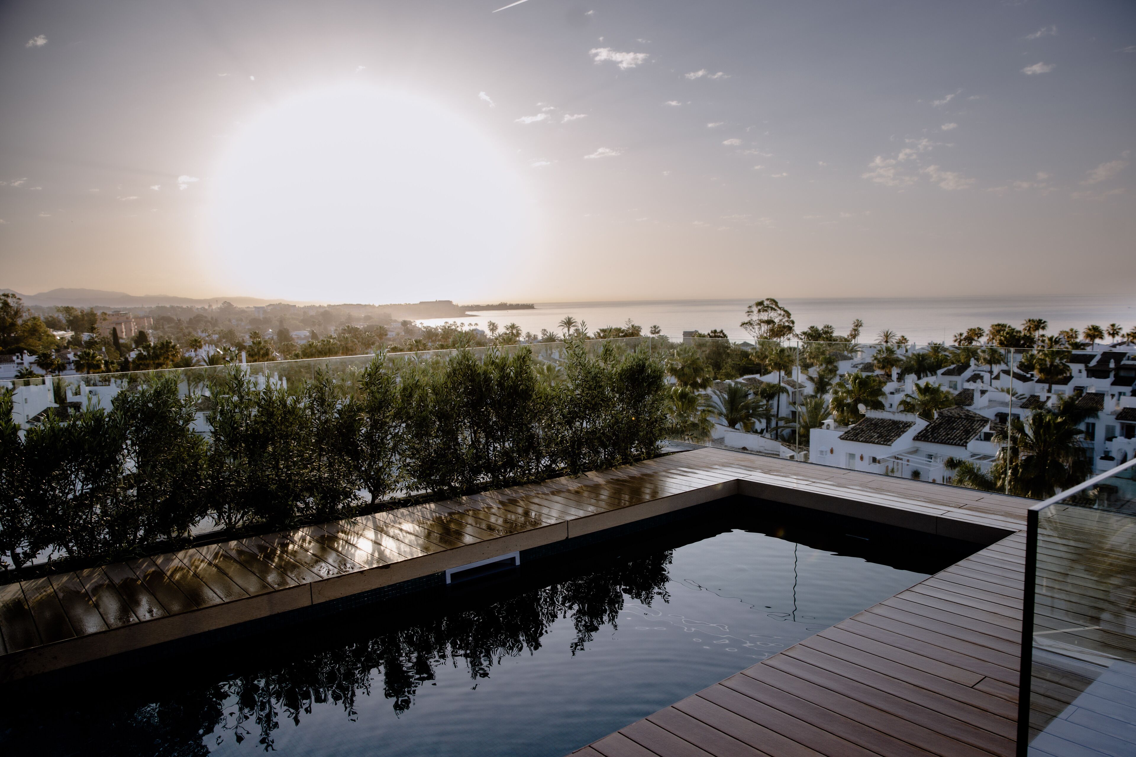 Una piscina infinita moderna con cubierta de madera, rodeada de vegetación exuberante y con vistas a una ciudad costera al atardecer.