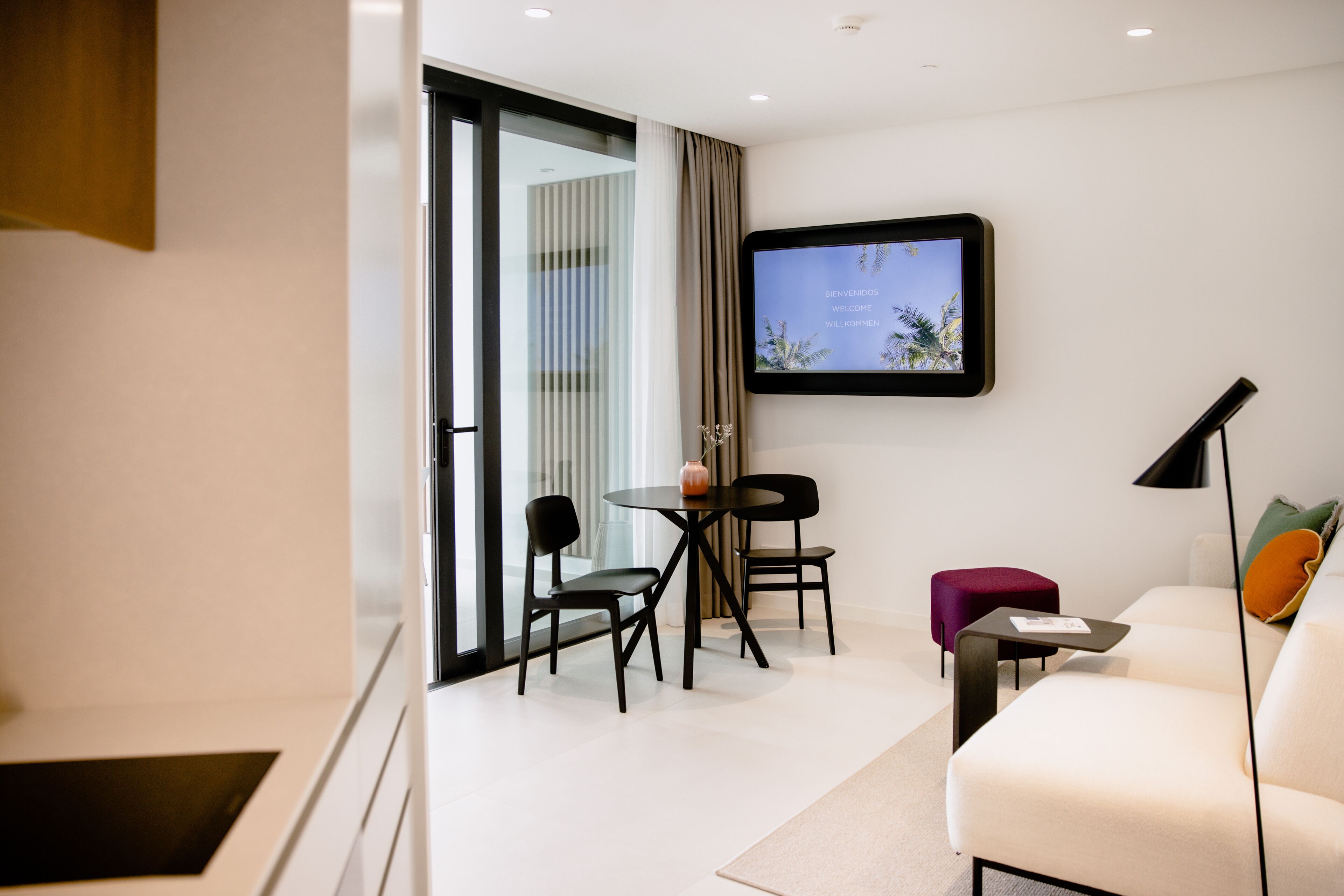 Ett modernt hotellrum med en soffa, ett litet bord, en TV som är monterad på väggen och en skjutdörr i glas som leder ut till en balkong.