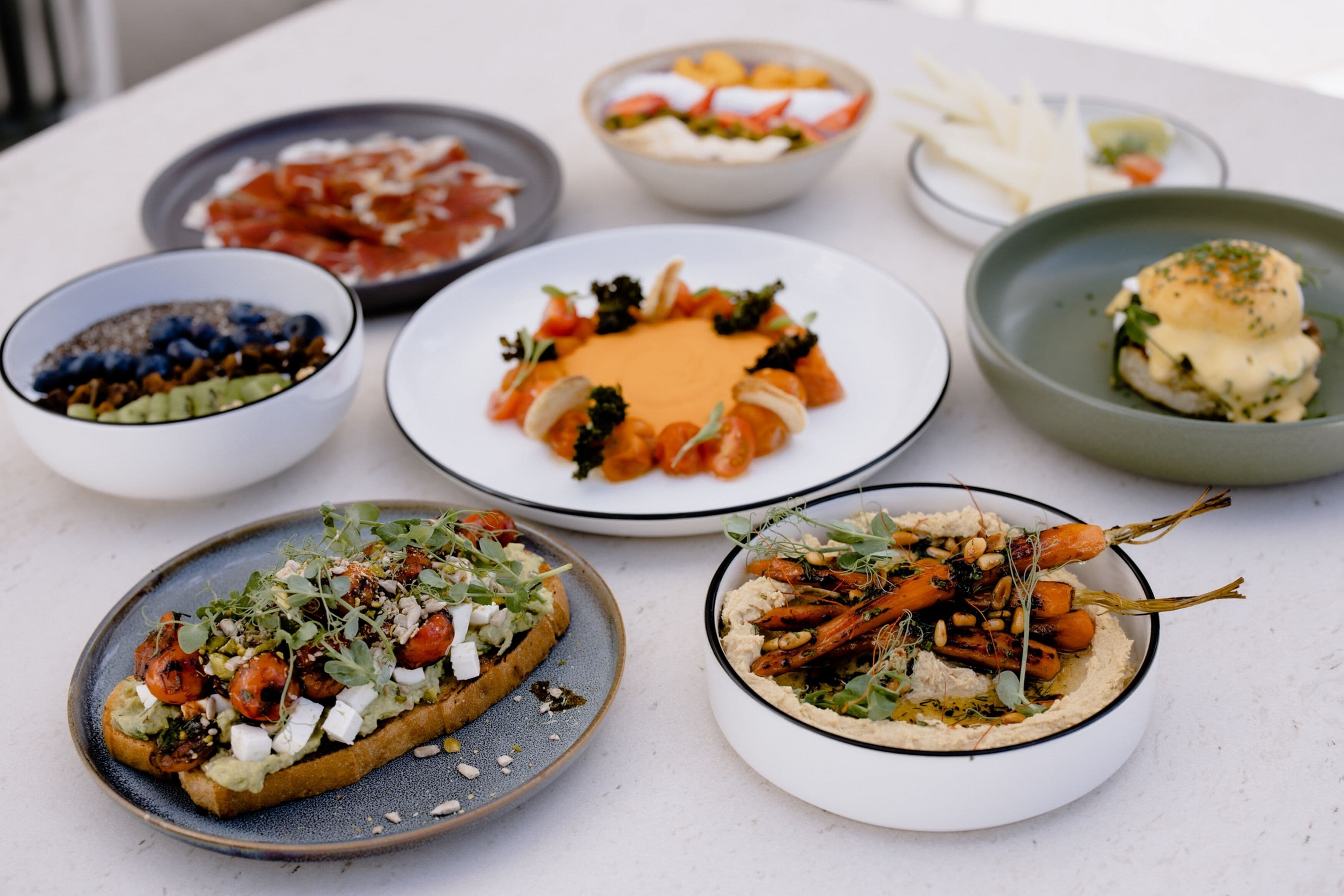 Forskellige retter på et bord, heriblandt en tallerken med fiskefilet, en skål med hummus og gulerødder, og en tallerken med brød overdækket med grøntsager og ost.
