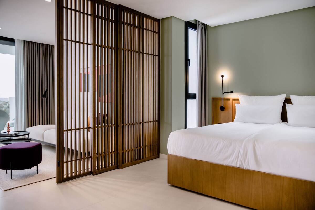 A modern hotel room with a large bed, a wooden partition, a desk with a chair, and a view of the outdoors.