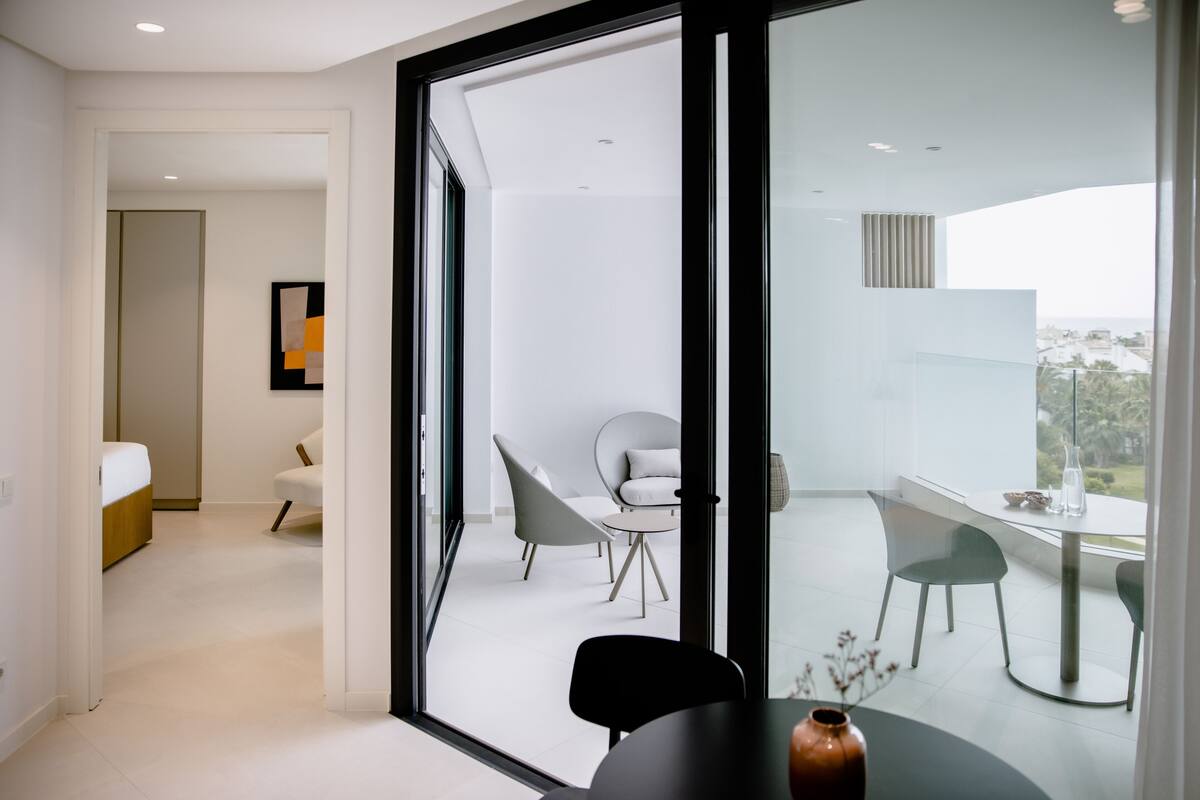 A modern hotel room with a balcony, a dining area, and a bed.