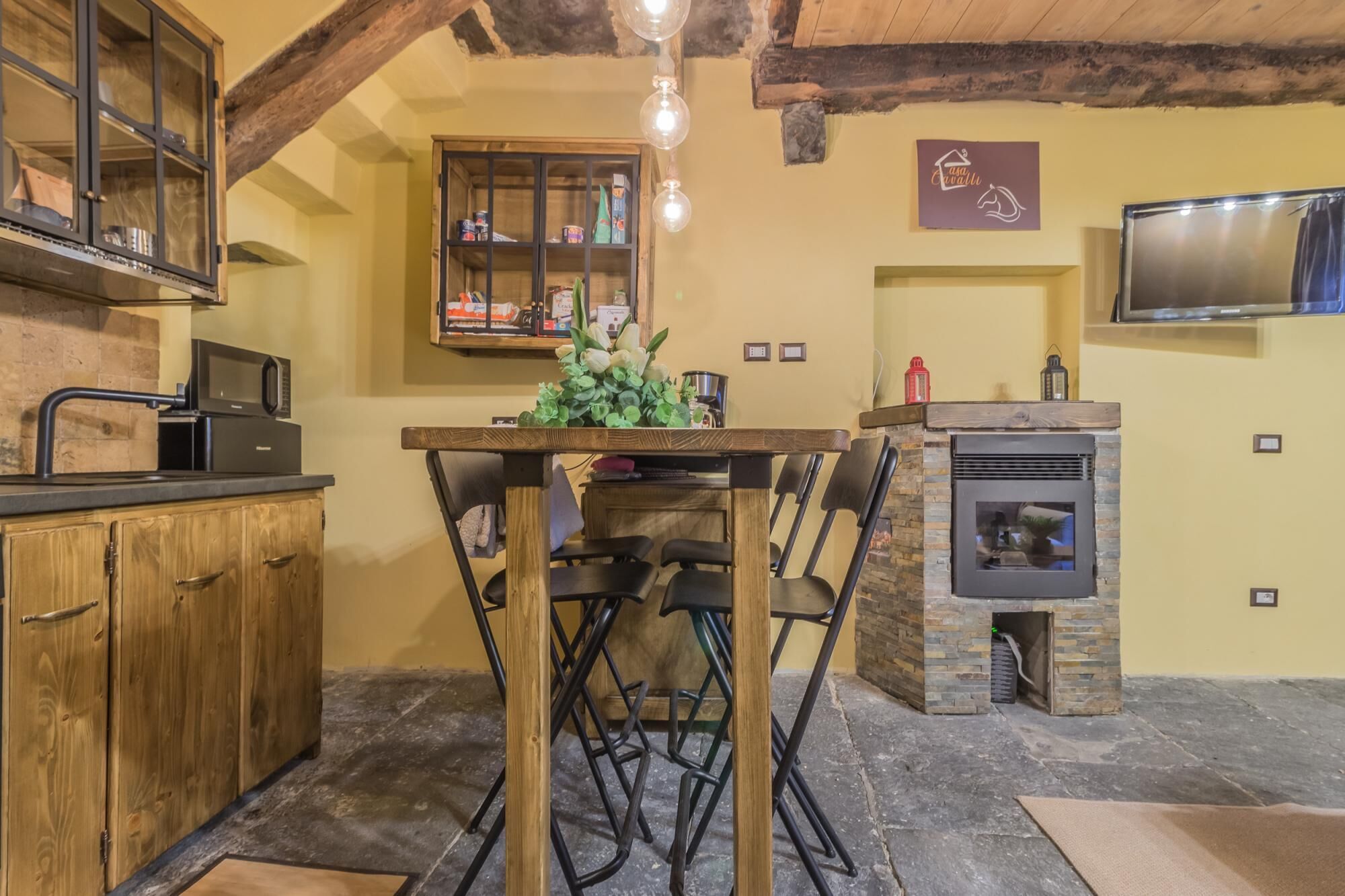A rustic kitchen with a wooden table, chairs, a stone fireplace, and a TV mounted on the wall.