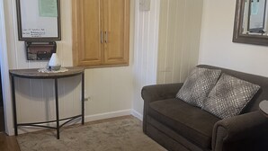 Une pièce avec une armoire en bois, une petite table avec une lampe, un canapé marron agrémenté d’un coussin à motifs et un tableau encadré accroché au mur.