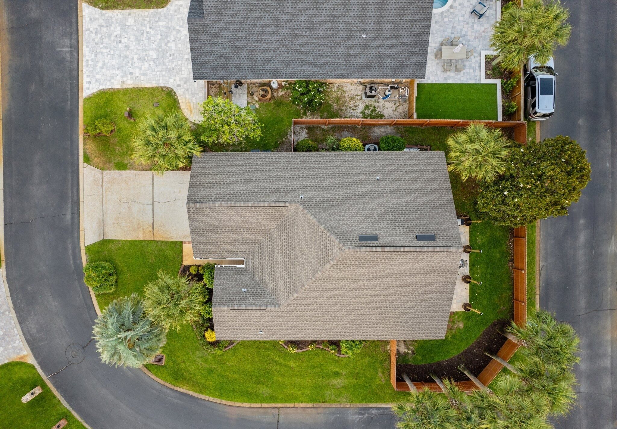 An aerial view of a residential property with a sloped roof, a driveway, and a well-maintained lawn with various trees and shrubs.
