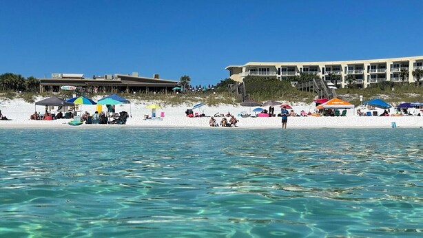 Een strand met helder water, parasols en mensen. Achtergrondgebouwen zijn zichtbaar.