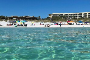 En strand med klart vand, parasoller og mennesker. Bygninger i baggrunden.
