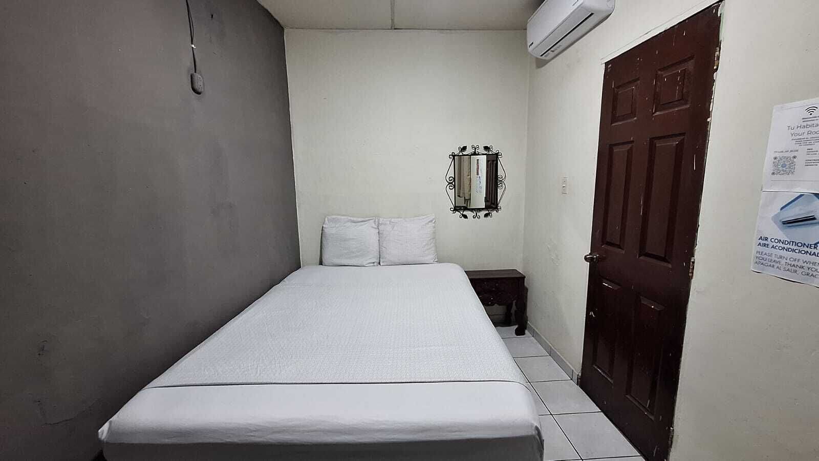 Una habitación pequeña y estrecha con una cama individual, una mesita de noche, un espejo en la pared y una puerta con un cartel.