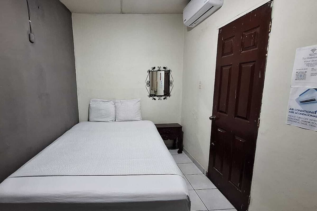 A small, narrow room with a single bed, a nightstand, a wall-mounted mirror, and a door with a posted sign.