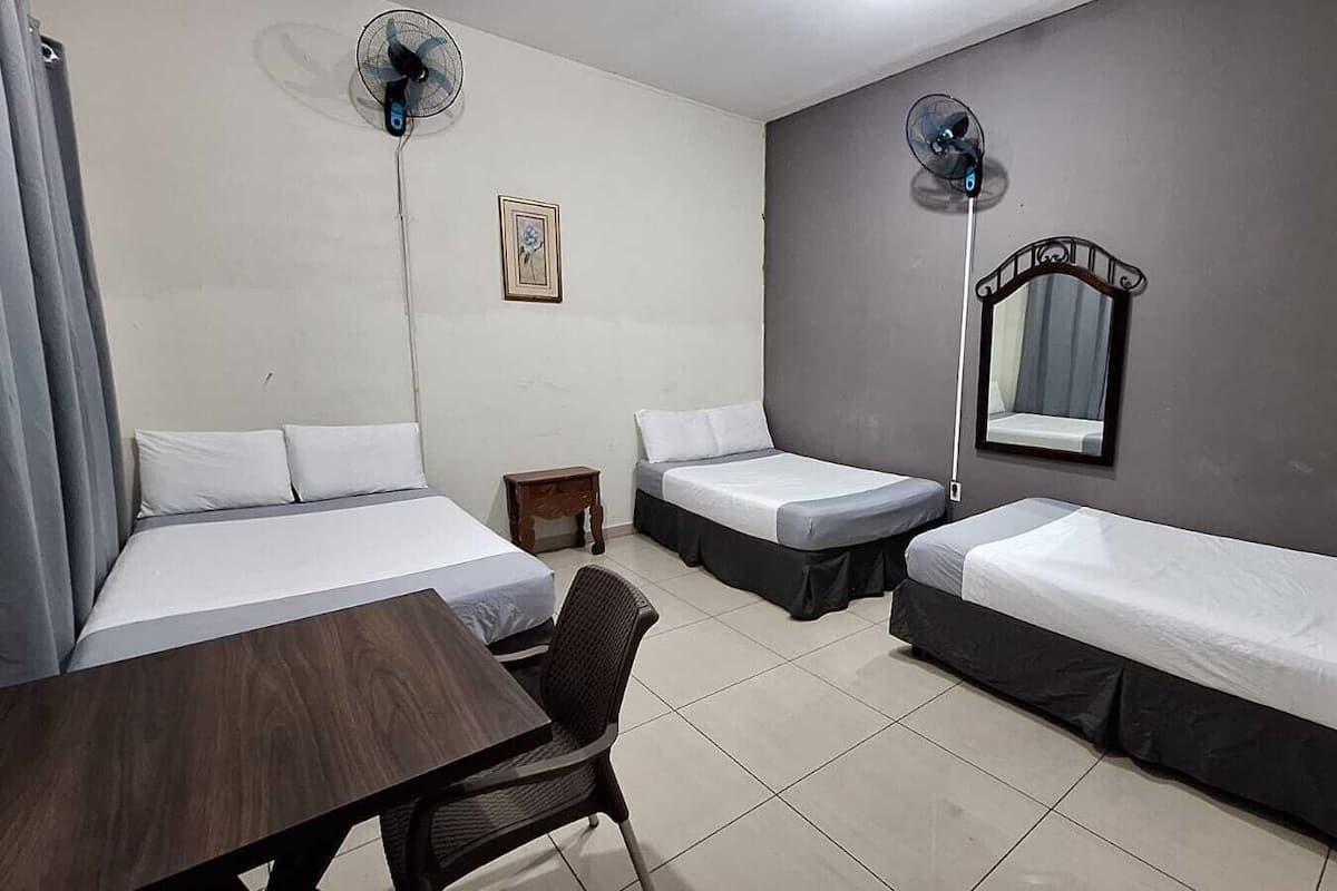 A hotel room with two single beds, a small table, a chair, two standing fans, and a mirror on the wall.