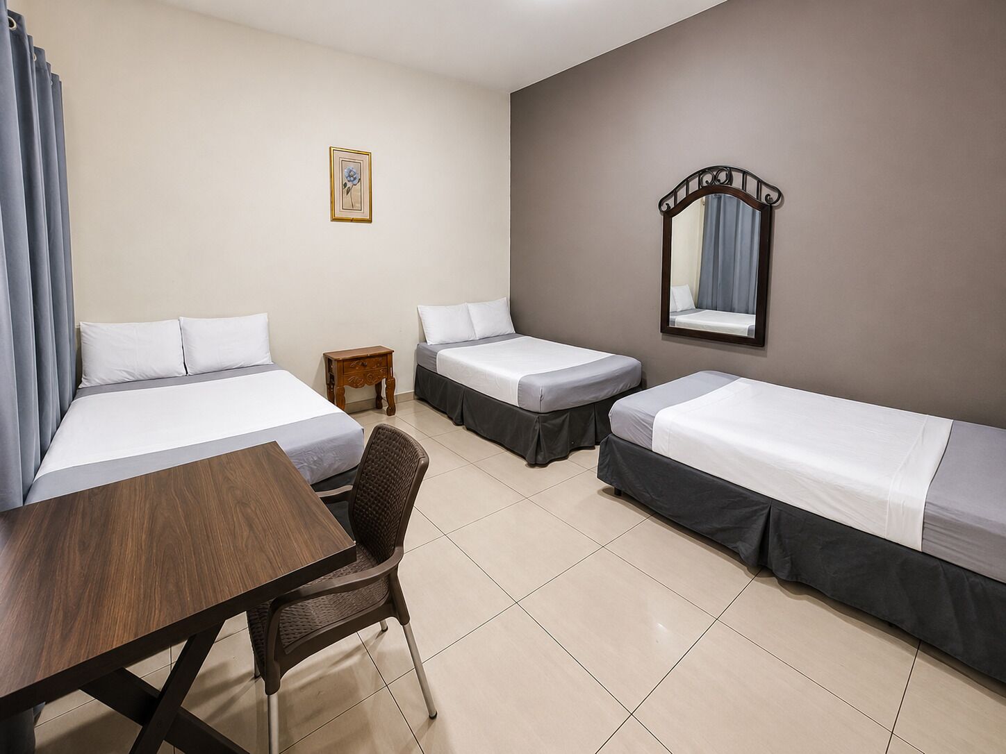 A hotel room with two single beds, a wooden desk, a chair, a small bedside table, a mirror, and a framed picture on the wall.