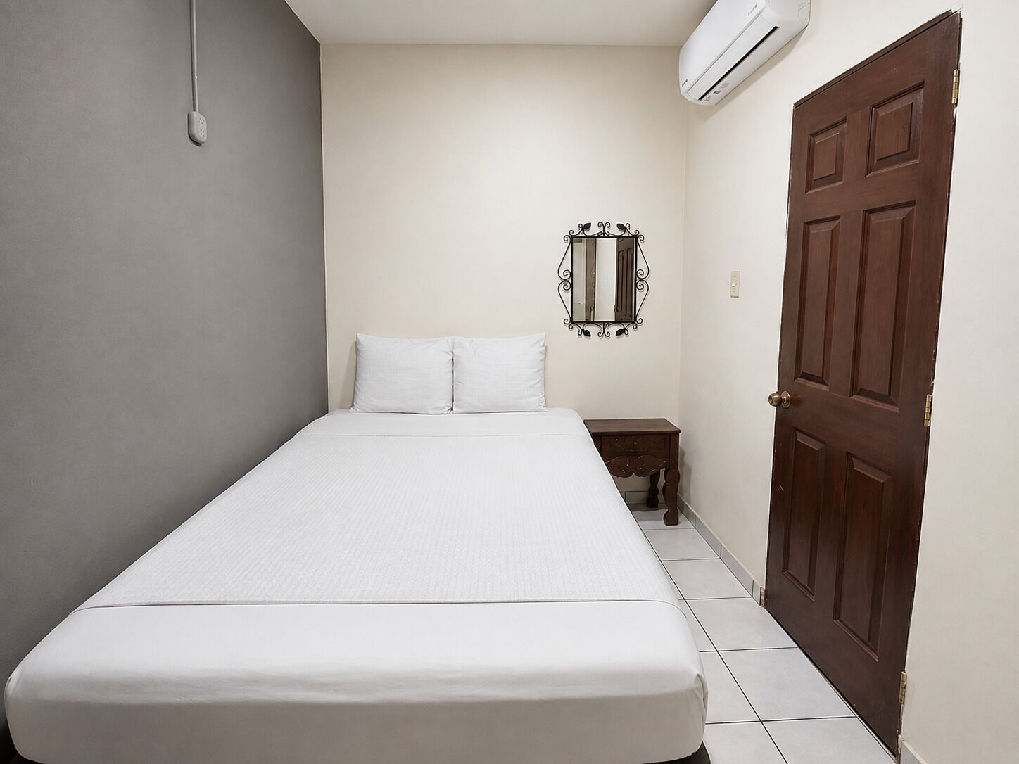 A small, single-bed room with a wooden door, a small bedside table, and a wall-mounted mirror.