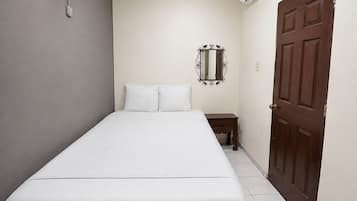 Ein kleines Zimmer mit einem Einzelbett, einer Holztür, einem kleinen Nachttisch und einem an der Wand befestigten Spiegel.