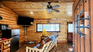 A log cabin interior with a wooden ceiling, a ceiling fan, a television mounted on the wall, a window with blinds, a wooden bench, and a refrigerator. - Music Mountain Retreat Cabin B (Hot Springs)