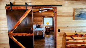 A wooden cabin interior with a bed, a window with a view, and a painting on the wall. - Music Mountain Retreat Cabin B (Hot Springs)