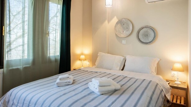 A neatly made bed with striped bedding, two wall-mounted decorative plates, a ceiling light, and an air conditioning unit. - Stagiates Guest House Room 2 (Volos)