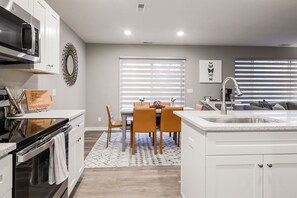 Una cocina moderna con gabinetes blancos, horno empotrado, microondas y una isla grande con fregadero. Hay una zona de comedor con mesa y sillas, y una ventana con persianas.
