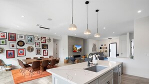 A modern kitchen with a marble island, pendant lighting, and a dining area with a wooden table and chairs. - Desert Escape | Poolside BBQs, Sunset Views & Spacious Comfort (Phoenix)