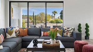 A modern living room with a sectional sofa, a coffee table with decorative items, and a view of the outdoors through large windows. - Desert Escape | Poolside BBQs, Sunset Views & Spacious Comfort (Phoenix)