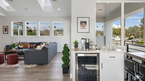 A modern living room with a grey sofa, a coffee table, and a small potted plant. There is a kitchen area with a white countertop and a built-in oven. - Desert Escape | Poolside BBQs, Sunset Views & Spacious Comfort (Phoenix)