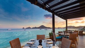 A dining area with tables set for two, overlooking a body of water with boats and a distant mountain range. - Villa del Arco Beach Resort & Spa Cabo San Lucas - All Inclusive (Cabo San Lucas)