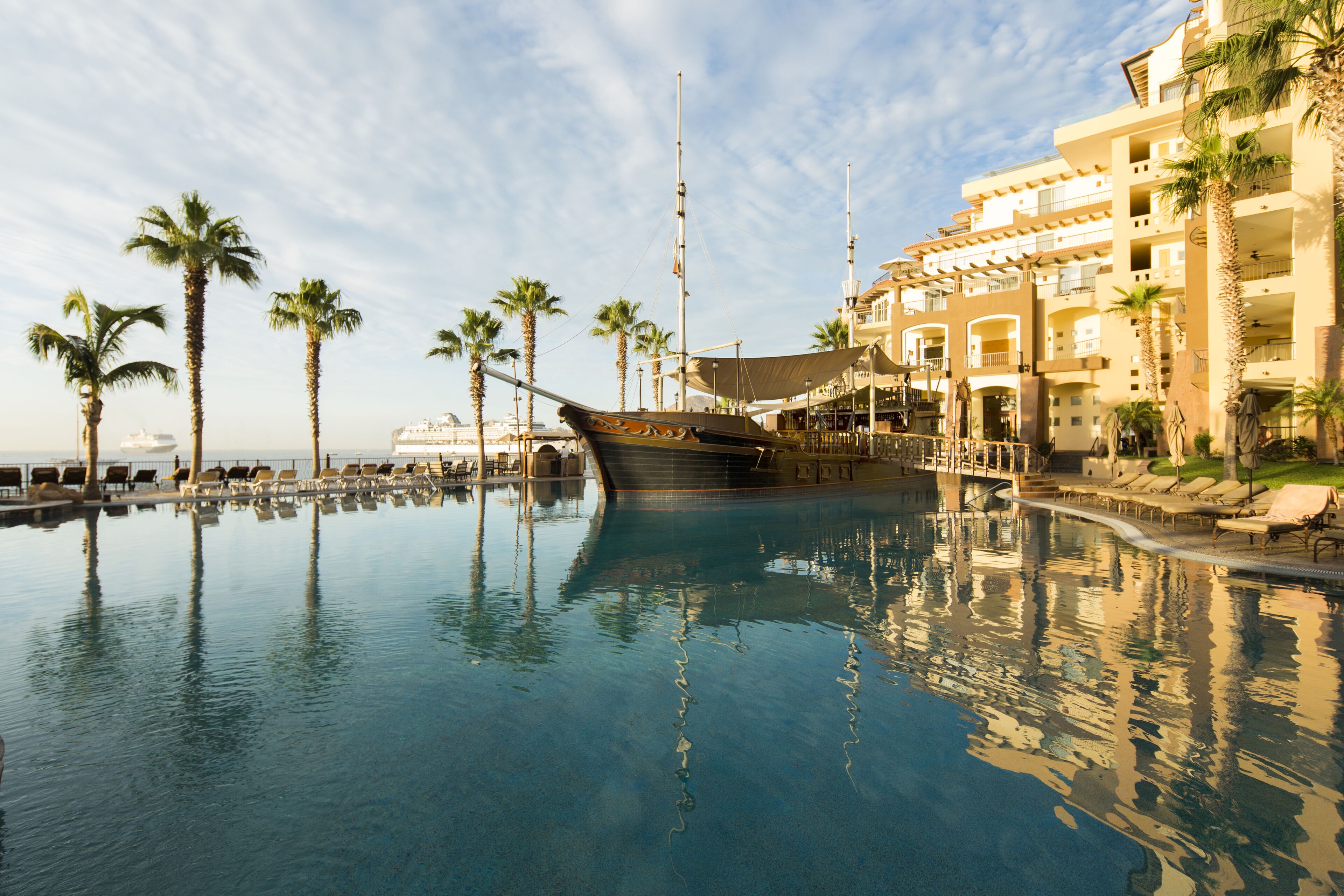 A large indoor swimming pool with a ship-shaped structure in the center, surrounded by palm trees and a multi-story building with balconies.