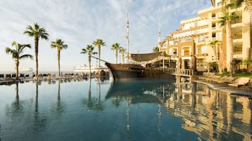 A large indoor swimming pool with a ship-shaped structure in the center, surrounded by palm trees and a multi-story building with balconies.