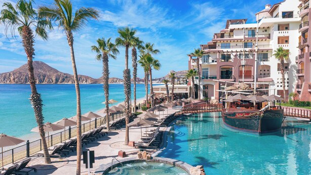 A resort pool area with lounge chairs and umbrellas, a boat, and a view of the sea and mountains. - Villa del Arco Beach Resort & Spa Cabo San Lucas - All Inclusive (Cabo San Lucas)