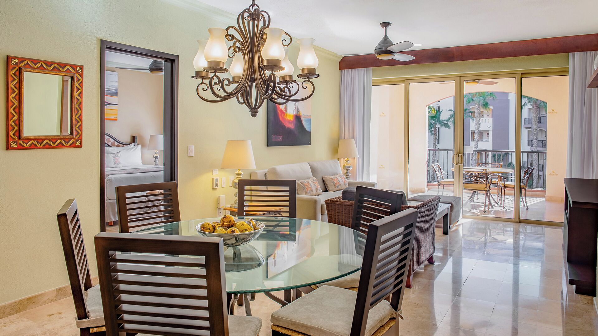 Un comedor con mesa de vidrio rodeada de sillas, una araña y vista del patio exterior.