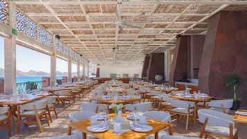 A spacious outdoor dining area with multiple tables and chairs, a ceiling with a woven pattern, and a view of the sea.
