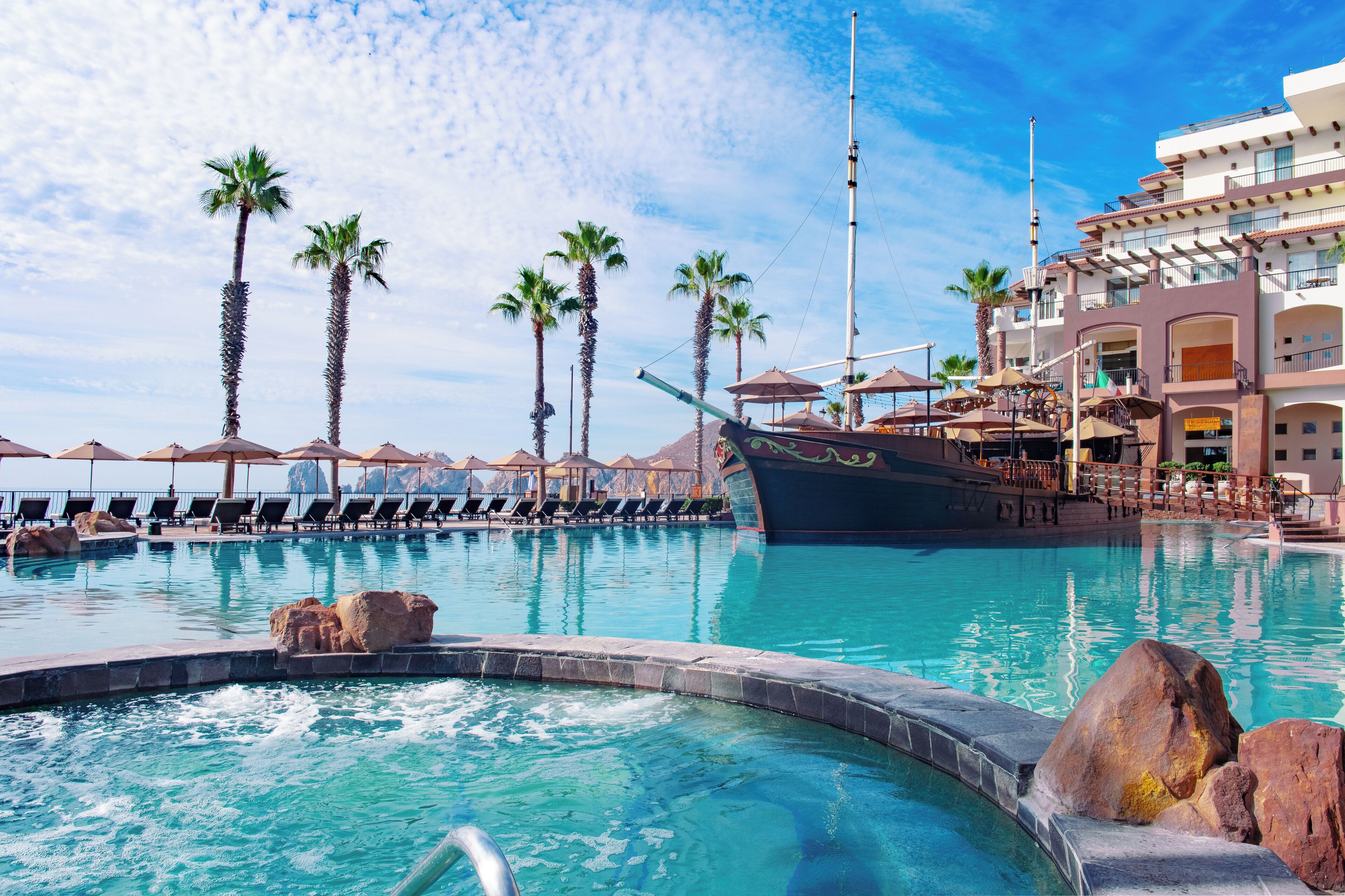 A pool area with lounge chairs and umbrellas, palm trees, and a large building with balconies.