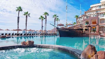 A pool area with lounge chairs and umbrellas, palm trees, and a large building with balconies.