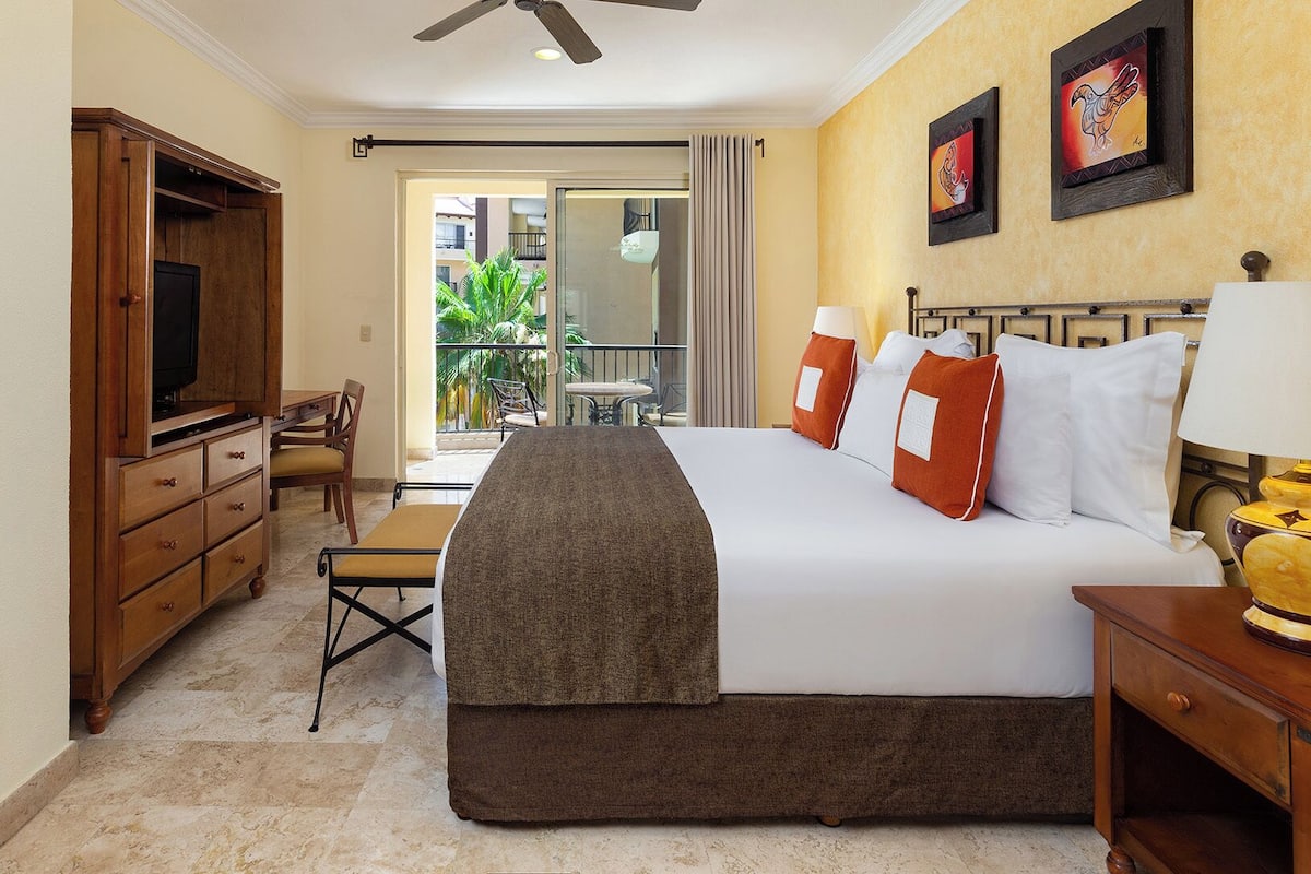 A hotel room with a bed, a desk, a TV, a ceiling fan, and a balcony with a view of palm trees.