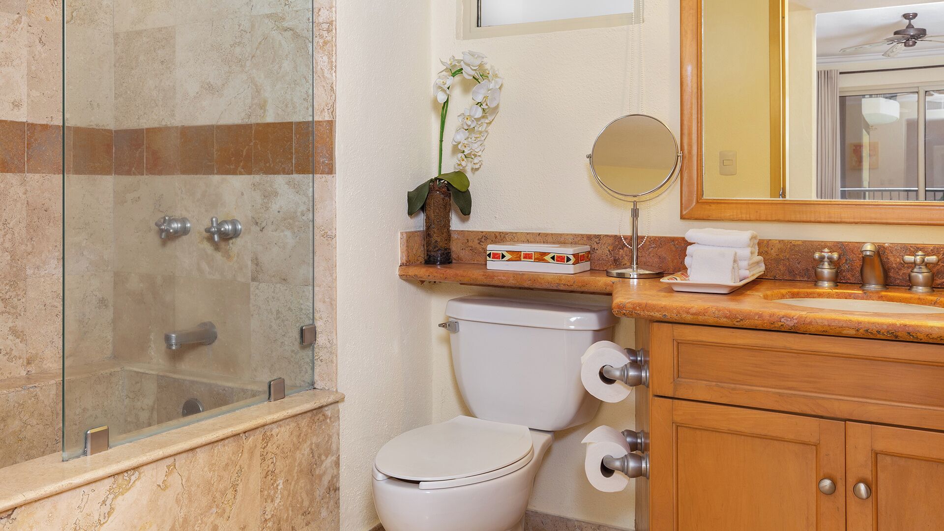 Un baño con cabina de ducha de vidrio, inodoro, lavabo con espejo y una orquídea decorativa.