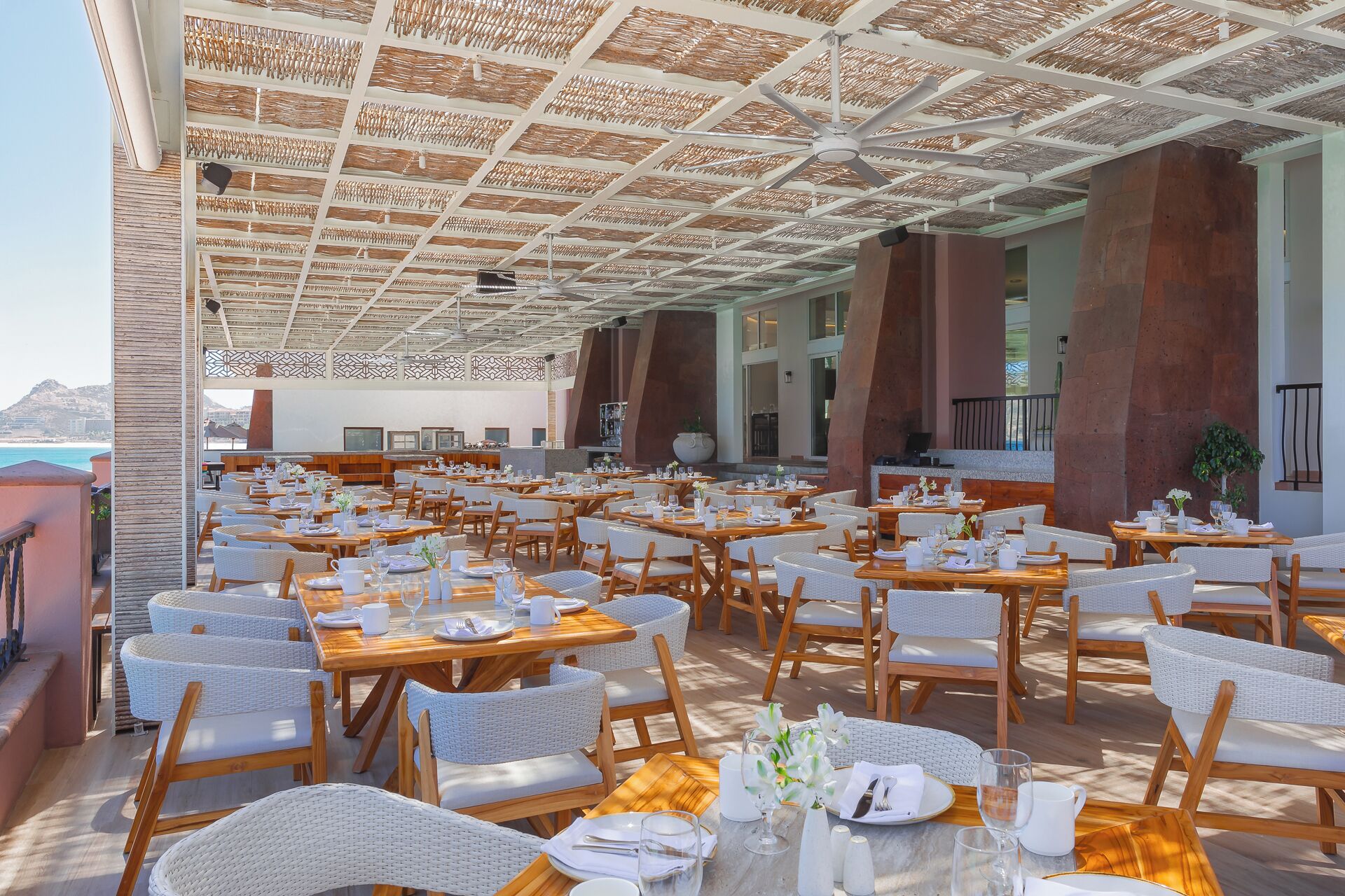 Un vaste espace extérieur aménagé pour les repas, avec de nombreuses tables et chaises, un plafond orné d’un motif tissé original et une vue sur l’océan.