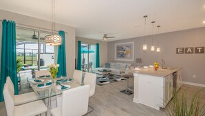 Eine moderne Küche mit Kochinsel, ein Essbereich mit Glastisch und eine Wohnfläche mit Sofa und Deckenventilator.